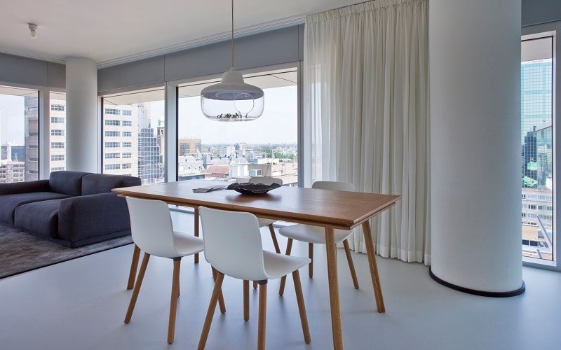 A modern dining area features a wooden table surrounded by three white chairs. Natural light enters through large windows dressed with sheer curtains, providing a view of the cityscape. A sleek pendant light hangs above the table, adding a contemporary touch to the space.