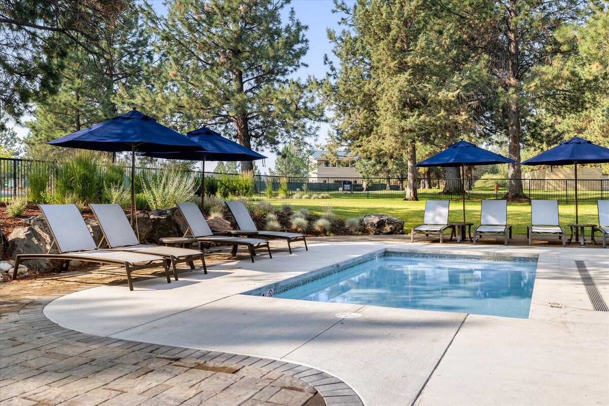 A community swimming pool is surrounded by lounge chairs and shaded by blue umbrellas. Lush greenery and tall pine trees border the pool area, providing a serene outdoor setting for relaxation. The pool features a smooth, curvy edge, inviting guests for a refreshing swim.