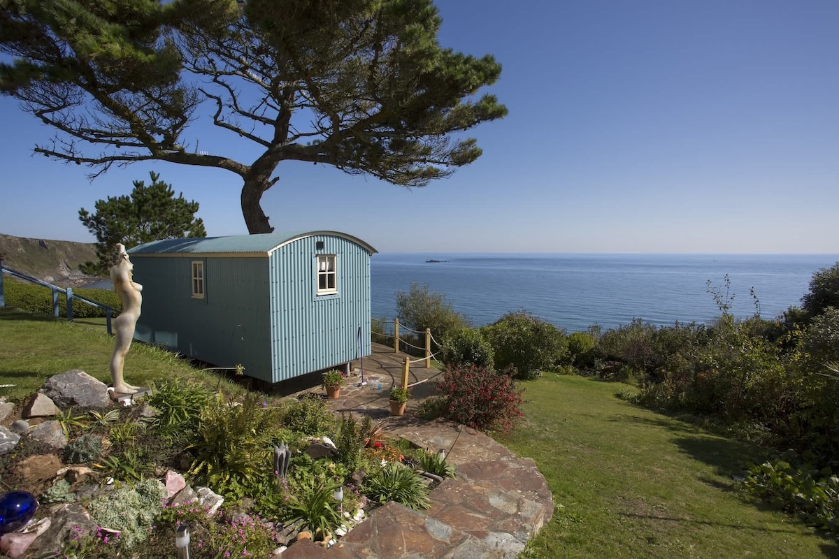 A charming beach-style shepherd's hut sits on a lush lawn with panoramic ocean views. The exterior features a light blue facade, and a large tree provides shade. Vibrant flowers and greenery surround the hut, creating a serene atmosphere, while the sea sparkles in the background.