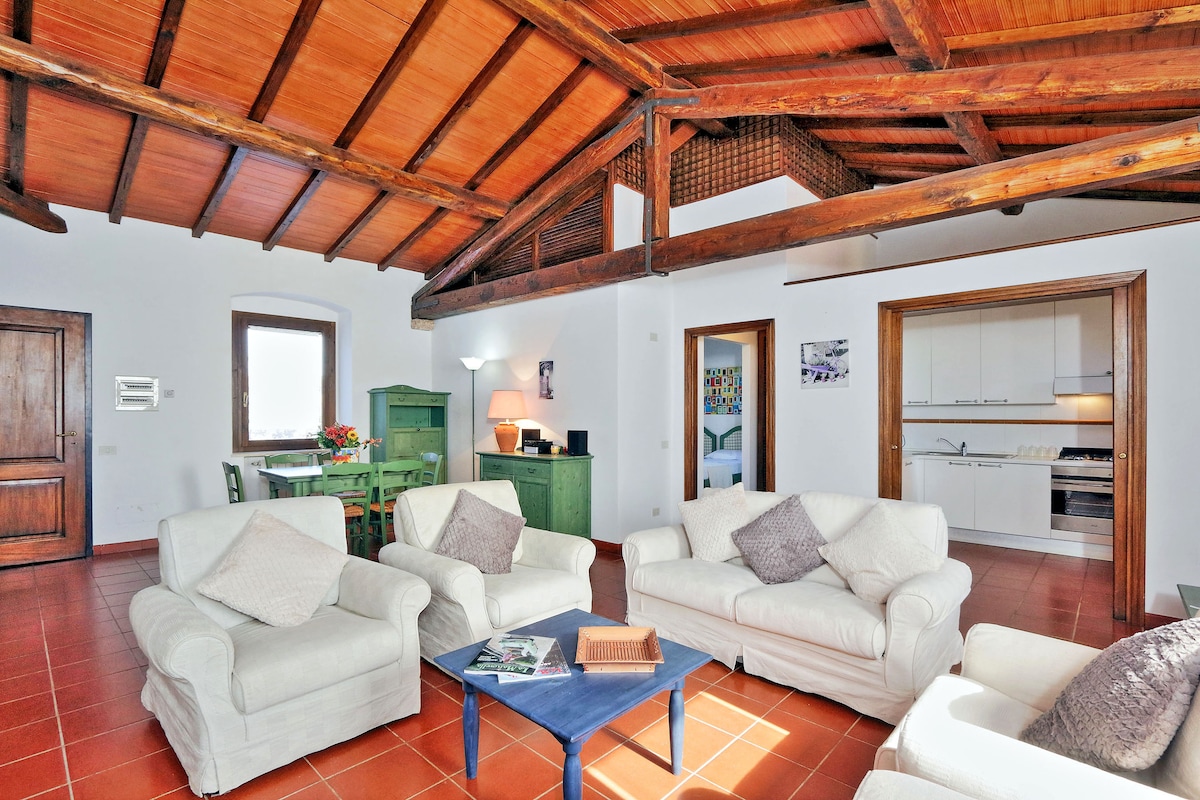 A spacious living area features high, wooden beam ceilings and terracotta tile flooring. Two white sofas and a blue coffee table are arranged in the center. A green wooden cabinet and dining table are visible, while a well-equipped kitchen is situated in the background.