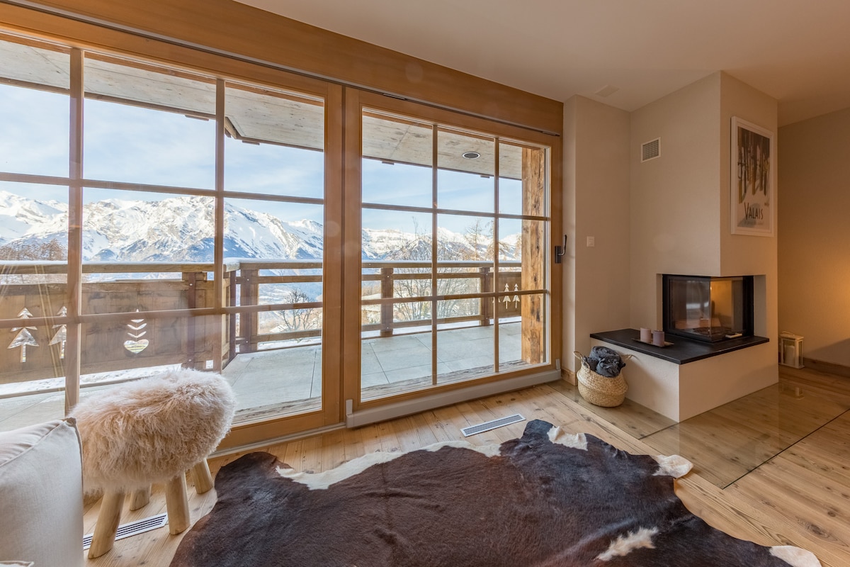 A living room area features large sliding glass doors that open to a spacious balcony with mountain views. A cozy area rug is positioned on the wooden floor, and a minimalist fireplace is integrated into the wall, enhancing the inviting aesthetic of the space.