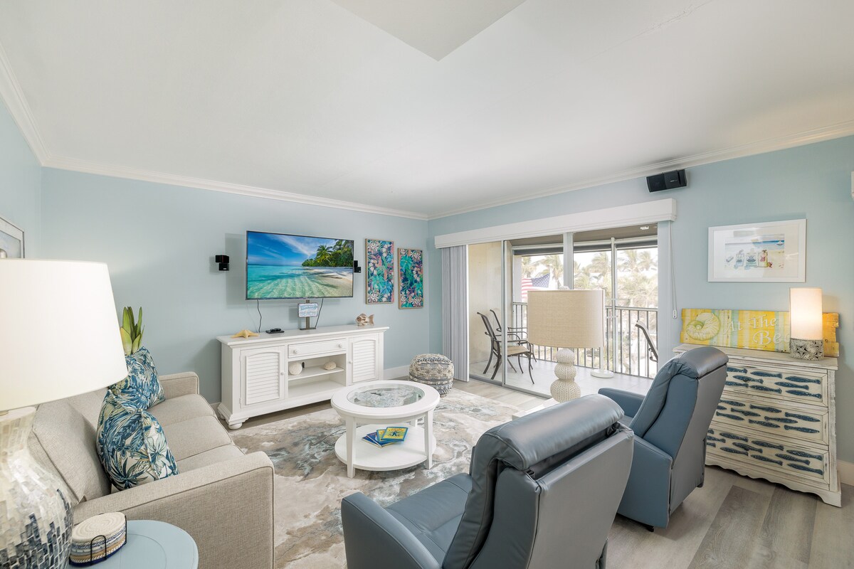A spacious living area features a light blue accent wall and tropical artwork. A comfortable gray sofa is paired with two recliners. A light-colored coffee table occupies the center, while a large TV is mounted on a white console. Natural light streams in from the sliding glass doors.