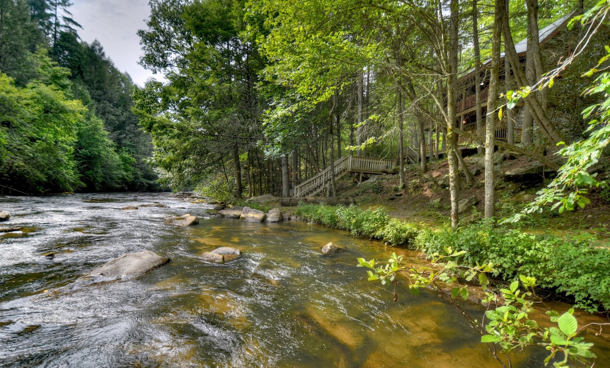 A scenic view showcases Fightingtown Creek with its gently flowing water and large natural rocks. The shaded area is framed by lush greenery and trees, while a set of wooden stairs leads up to the cabin nestled among the foliage.