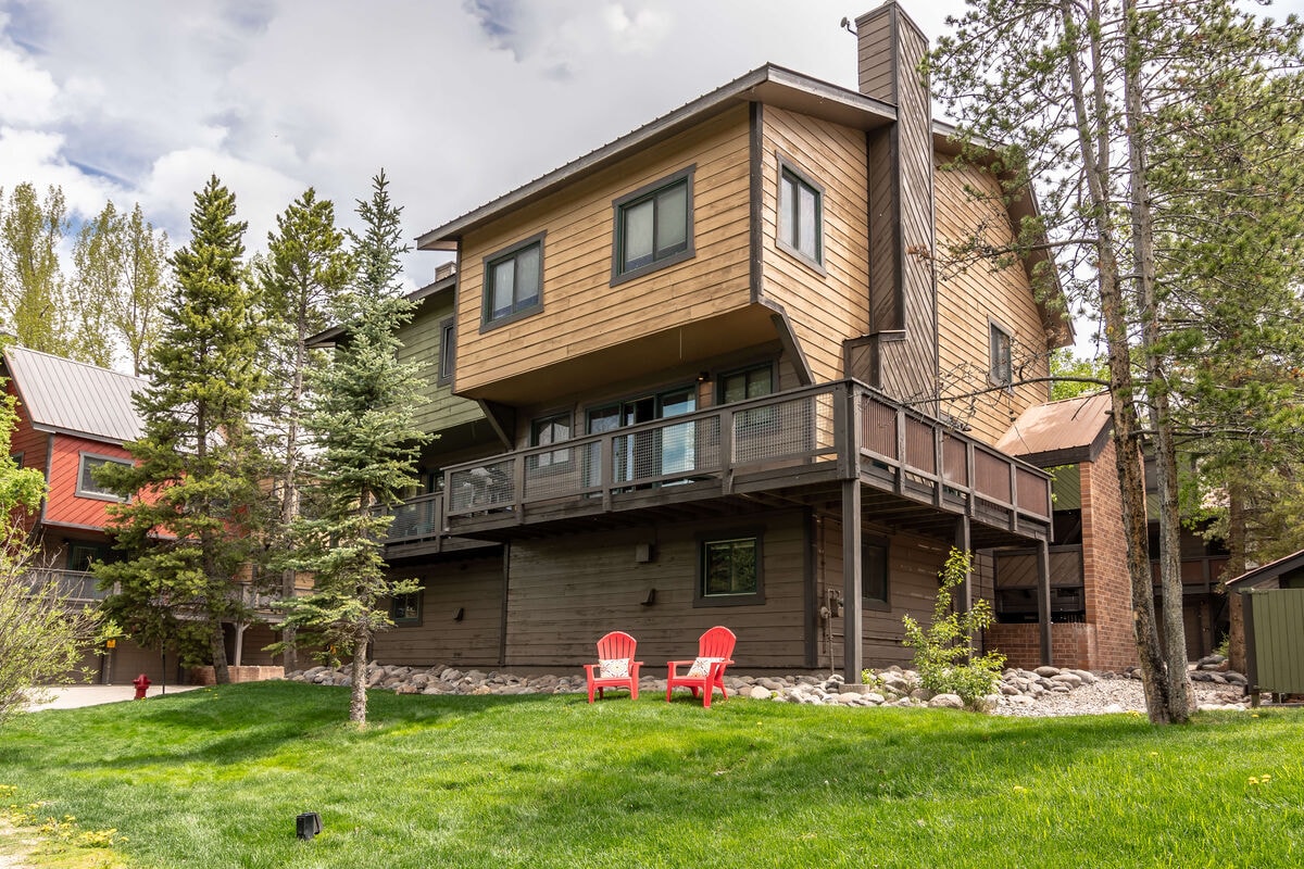 A three-level townhome features a wooden exterior and a large wrap-around balcony surrounded by landscaped greenery. Two red Adirondack chairs are positioned on the lawn, providing a comfortable spot to relax. Surrounding trees add natural beauty to the inviting outdoor space.