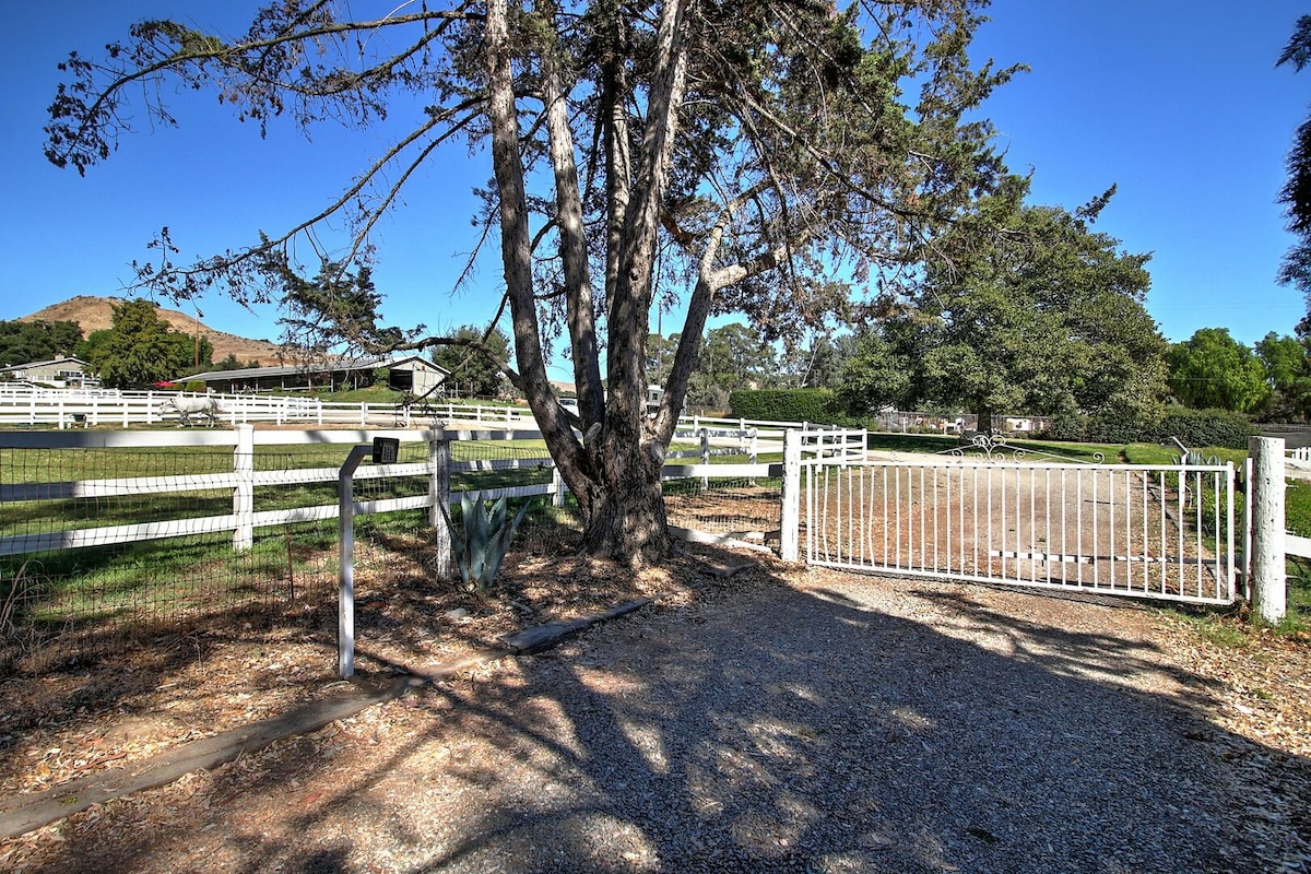 A white metal gate stands open, leading into a spacious ranch area. Shadows of a large tree stretch across the gravel driveway, while lush green pastures and a white fence define the property boundaries. The distant hills are visible under a clear blue sky.