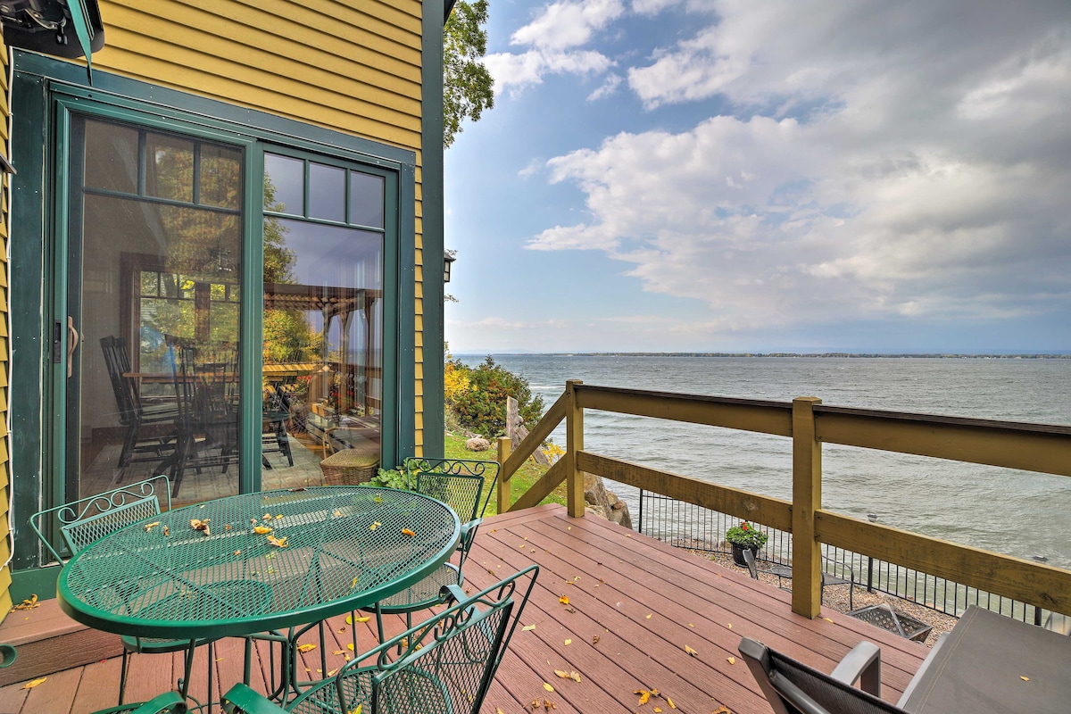 A spacious deck is visible, furnished with a green bistro table and chairs. The deck overlooks the expansive Lake Champlain, with rippling water reflecting the sky. Large glass doors provide access to the indoor living area, blending the outdoor and indoor settings.