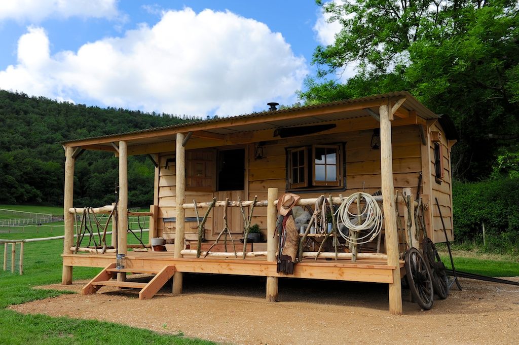 A wooden cabin is set on a gravel area, featuring a covered porch with rustic railings. Various ropes and tools are displayed on the porch. Lush greenery and a distant wooded area are visible in the background under a partly cloudy sky.