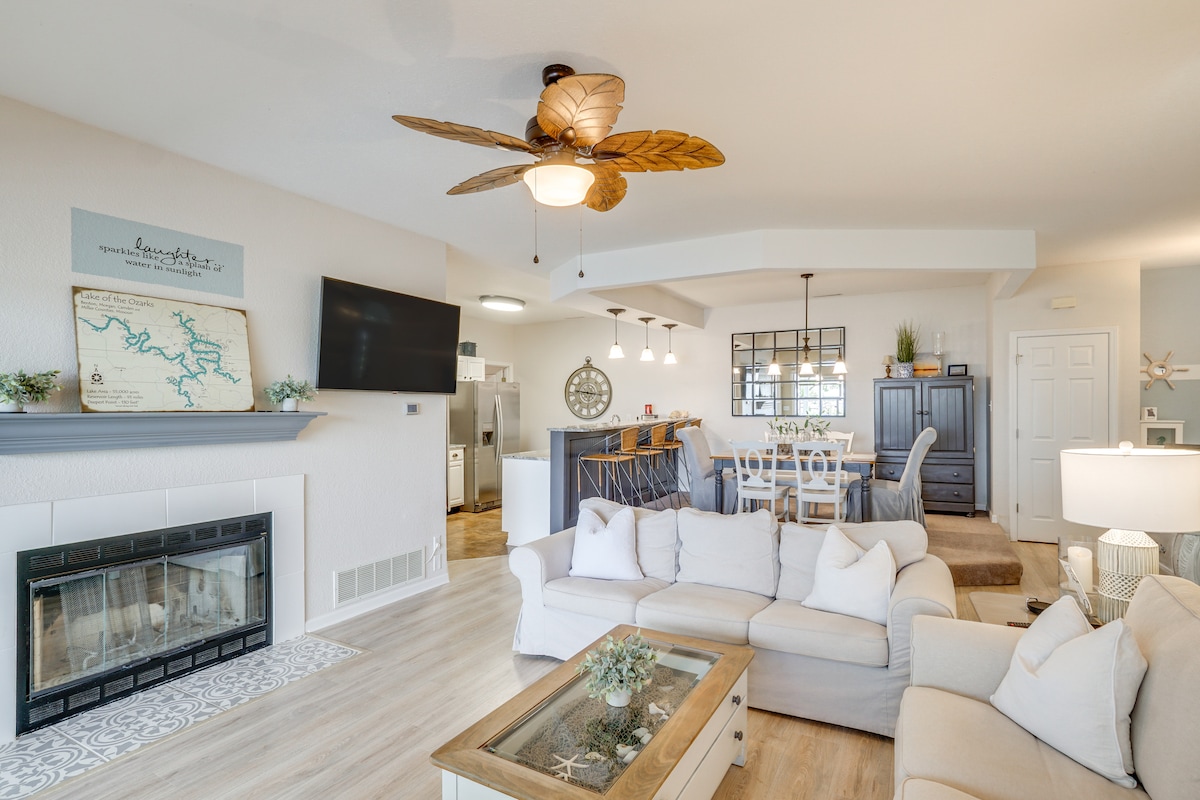 A combined living and dining area features a cozy layout with a sectional sofa and a coffee table centered around a wood-burning fireplace. A large flat-screen TV is mounted on the wall, and a dining table with chairs is visible in the background, accentuated by natural light.