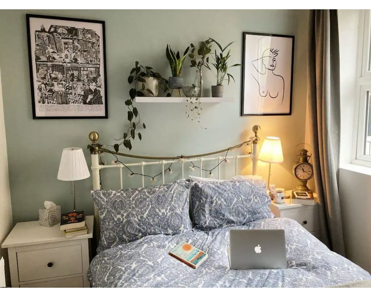 A cozy double bedroom is featured, with a patterned blue duvet and multiple pillows on the bed. Two framed artworks are displayed above the headboard. A small shelf hosts several plants, while bedside lamps provide soft illumination. A laptop rests on the bed beside an open book.