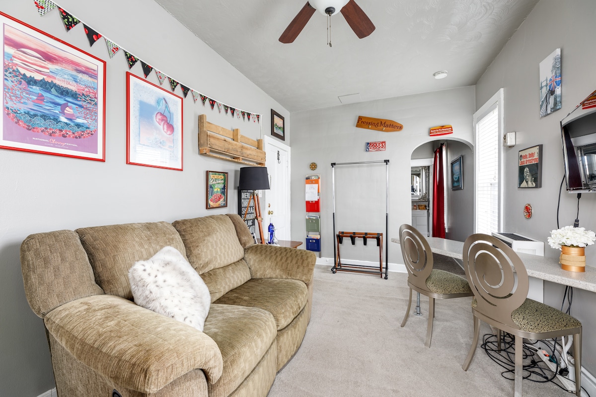 A spacious living area features plush seating accompanied by decorative wall art and playful bunting. A flat-screen TV is mounted nearby, while a small office desk with two stylish chairs offers a functional workspace. Natural light filters in through a window, enhancing the inviting atmosphere.