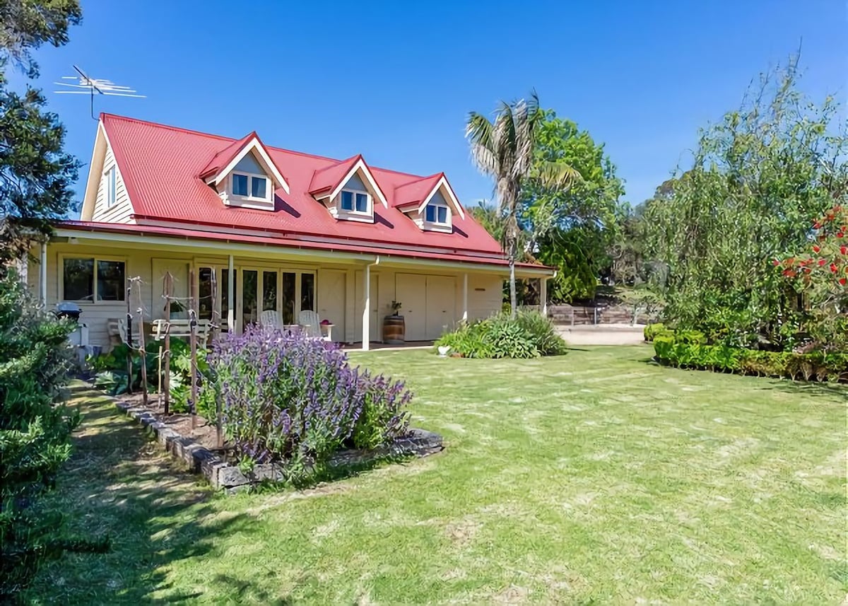 The exterior of a large, two-story property is showcased, featuring a red roof and multiple gabled windows. A spacious, manicured lawn is surrounded by vibrant flower beds, with several trees providing shade, creating an inviting garden space.