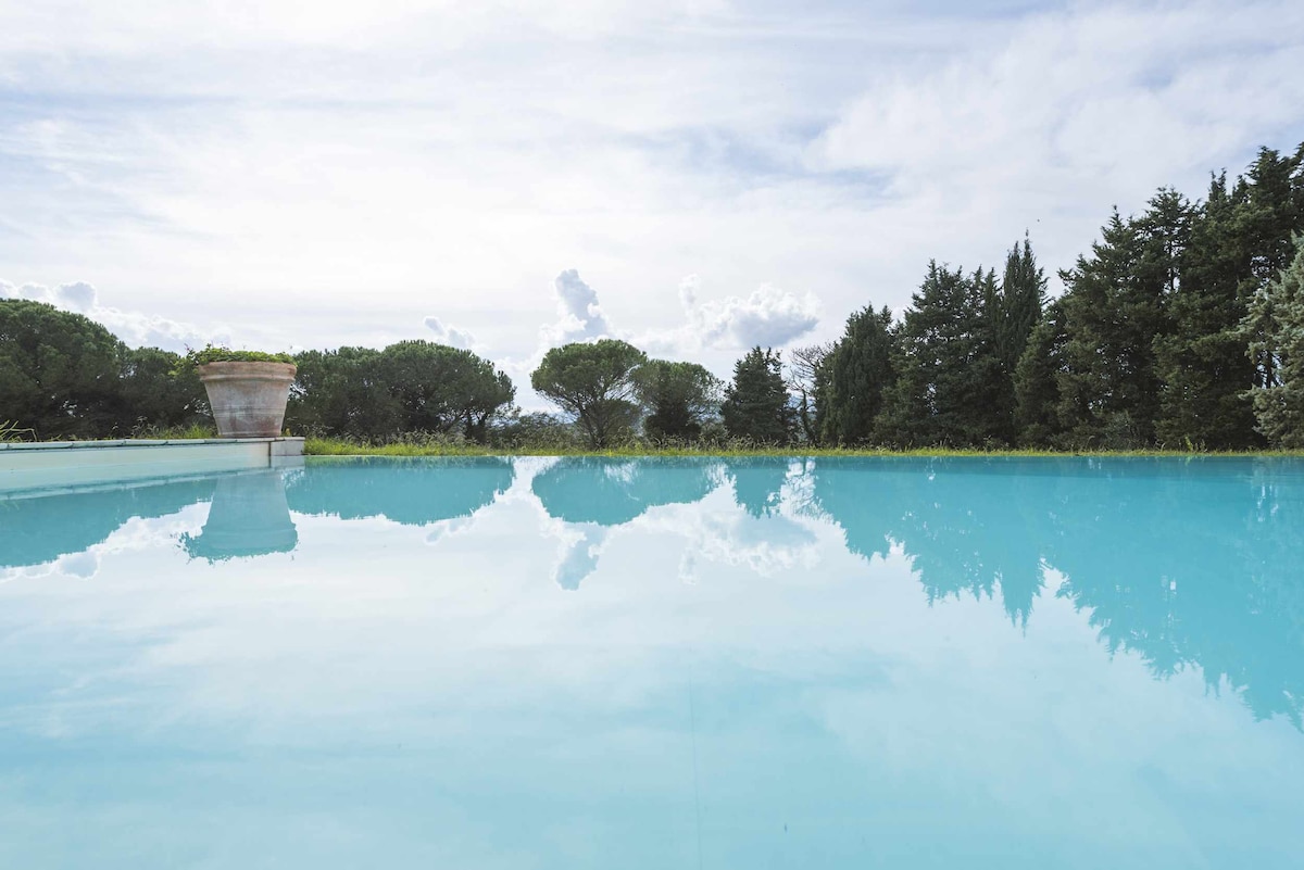 A tranquil infinity pool reflects the clear sky, with lush greenery framing the serene setting. The water appears invitingly blue, creating a harmonious connection between the natural landscape and the pool's edge.