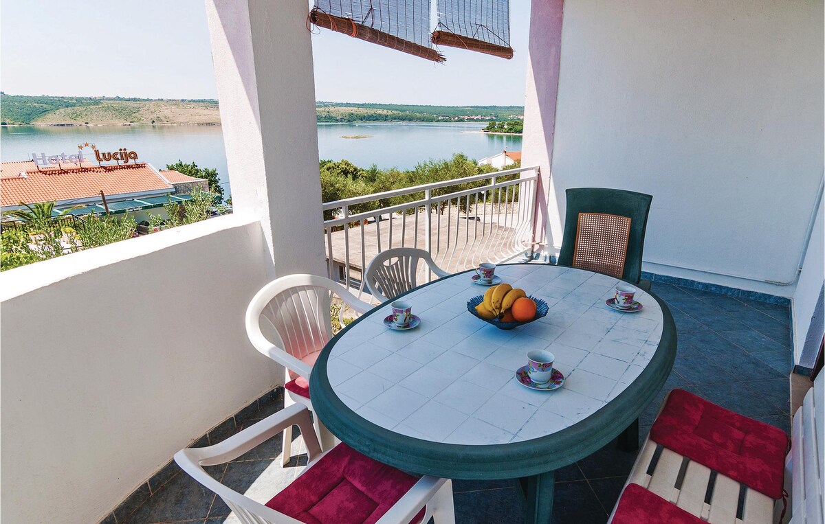 An outdoor seating area features a round table surrounded by chairs in light colors. Fresh fruits are displayed on the table, and a scenic view of the nearby water can be seen in the background. Sunlight illuminates the space, enhancing the tranquil atmosphere.