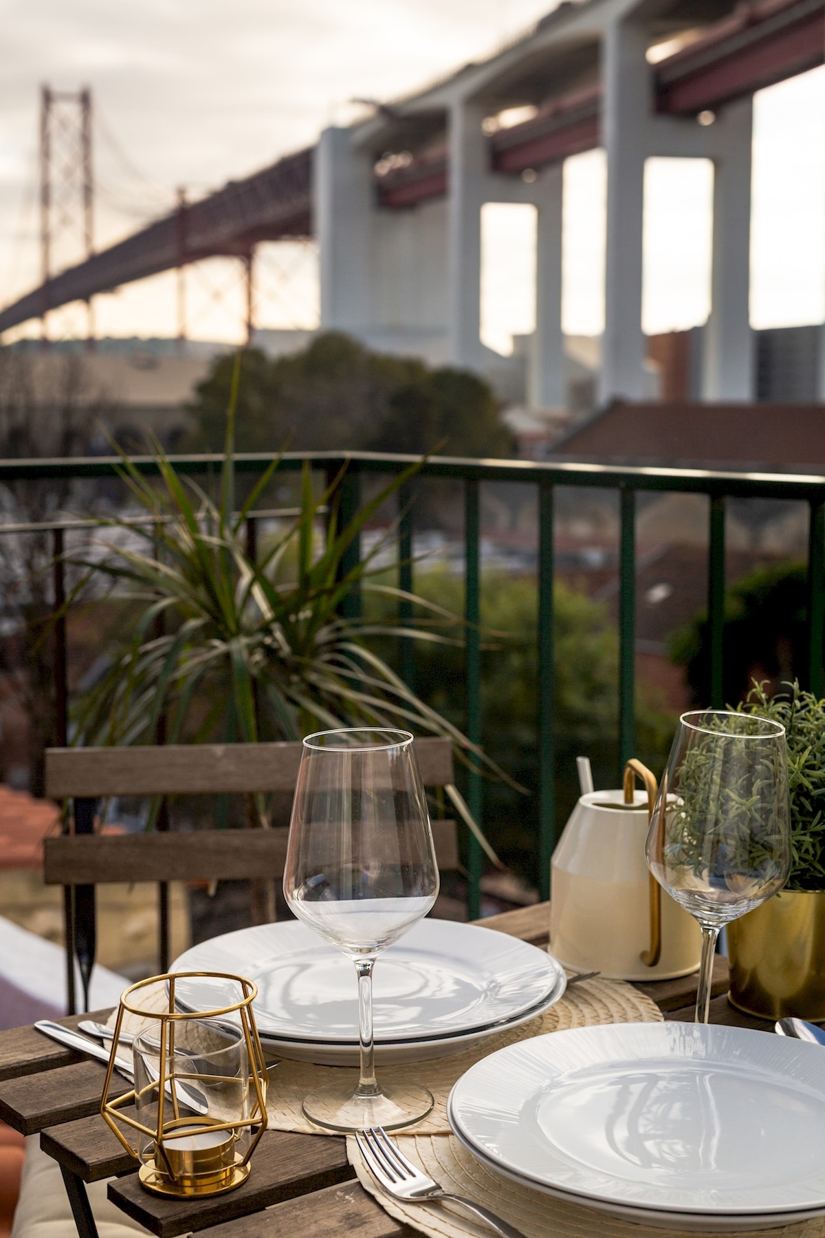 A dining setup is showcased on a terrace, featuring plates and glasses arranged on a wooden table. The background highlights a large bridge, complemented by greenery in view, adding to the outdoor ambiance.