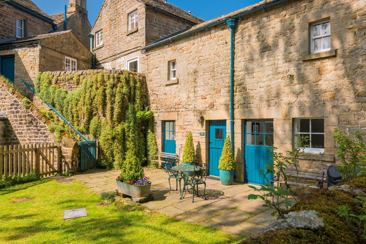 The exterior of the cottage presents a charming stone facade with bright blue doors, framed by lush greenery. A paved terrace area is furnished with a small table and chairs, inviting outdoor relaxation amidst garden plants. Steps lead to an upper level, enhancing the picturesque setting.