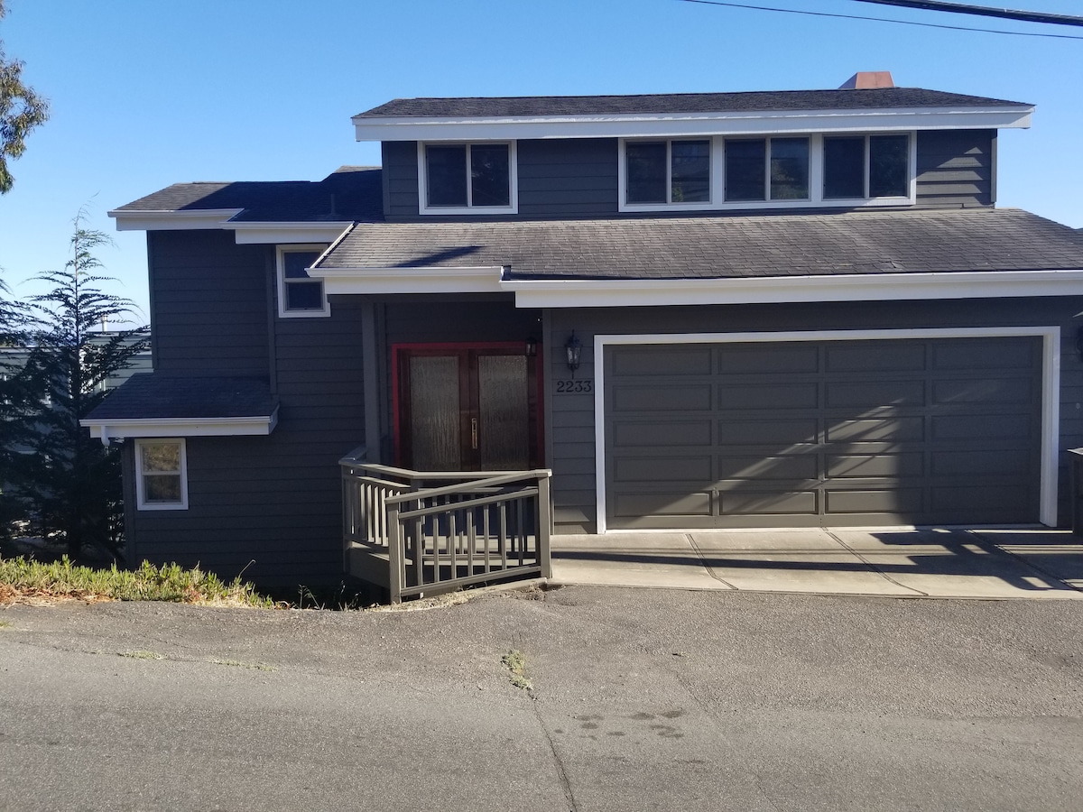The contemporary exterior of the vacation rental showcases a dark wood facade with large windows and a welcoming red front door. A driveway leads to a two-car garage. Surrounding greenery complements the design, while the home is positioned on a slight incline for enhanced views.