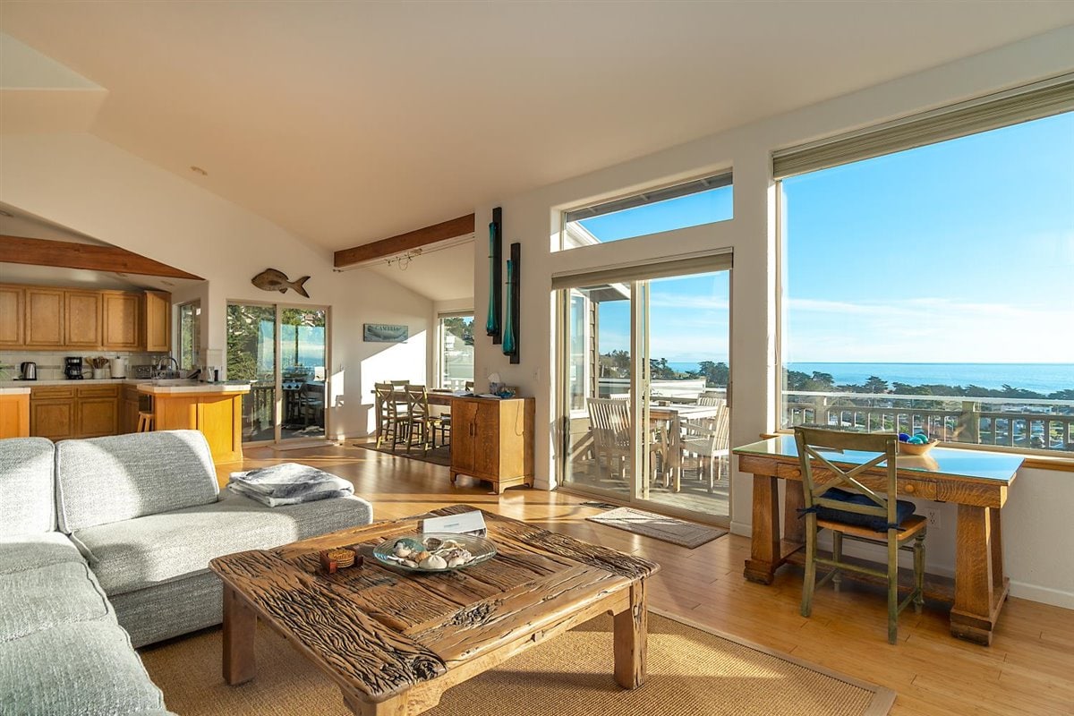 An airy living room features a spacious gray sofa and a rustic coffee table with decorative items. Large windows offer abundant natural light and stunning ocean views. The open layout connects to a dining area and a kitchen, enhancing the home's inviting atmosphere.