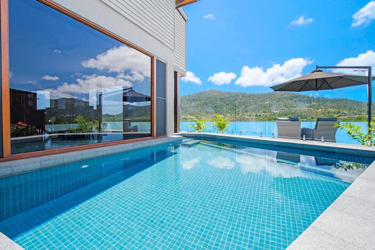 A sparkling pool, framed by sleek stone tiles, reflects the clear blue sky and surrounding greenery. Comfortable seating is positioned nearby under a shaded umbrella, offering a perfect spot for relaxation while enjoying the scenic waterfront views.