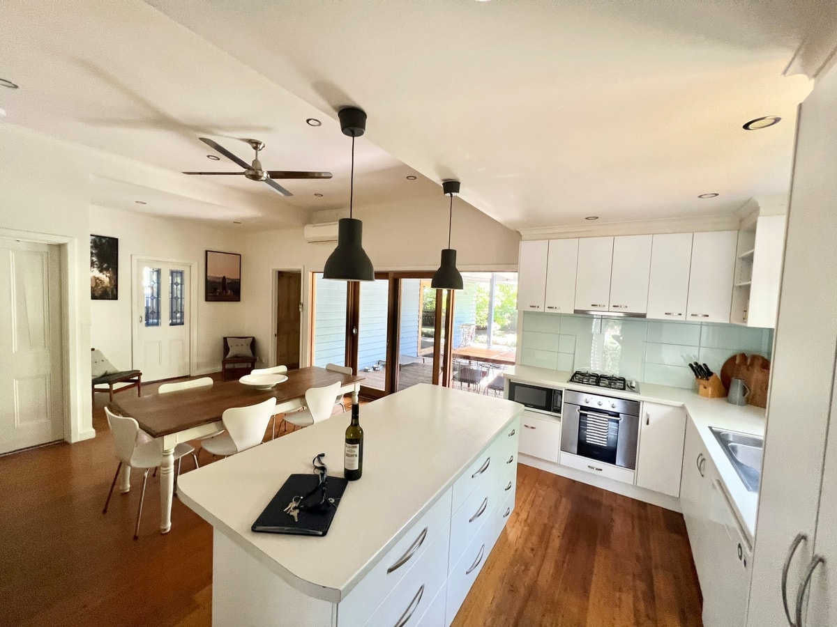 The open plan kitchen features a large island with storage, sleek cabinetry, and modern appliances. A dining table with white chairs is visible, while natural light floods the area through glass doors that lead to an outdoor deck.