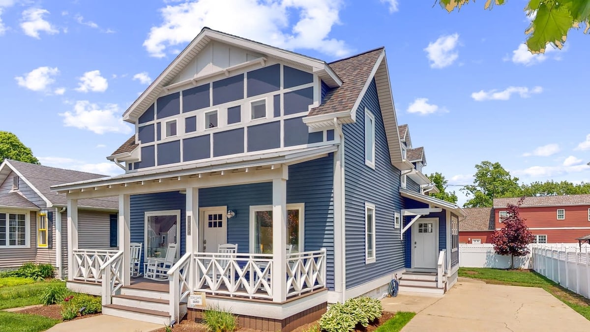 A charming two-story house features blue siding with white trim, showcasing a spacious front porch with decorative railings. The landscape is well-maintained, and the driveway leads to the entrance. Neighboring homes are visible in the background, contributing to a welcoming neighborhood setting.
