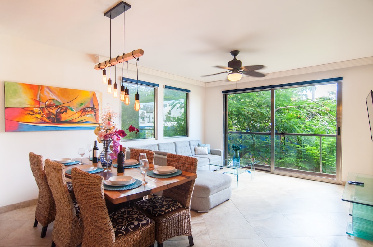 A comfortable dining area features a wooden table surrounded by six wicker chairs, set for a meal with plates and glasses. A stylish sectional sofa is visible, complemented by large windows that provide natural light and a view of lush greenery. Vibrant artwork decorates one wall.