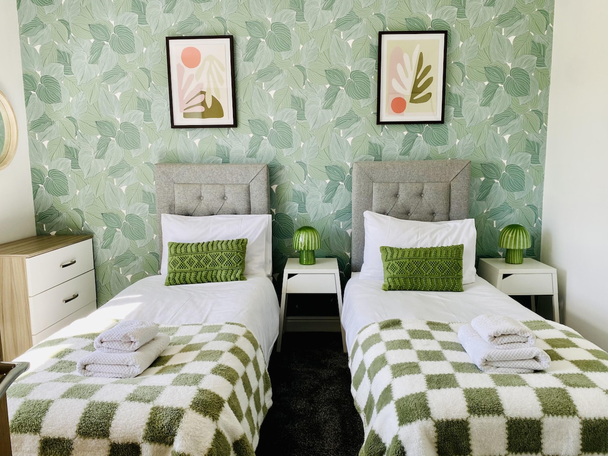 A bright bedroom features two single beds dressed in white linens, positioned against a green leafy wallpaper. Each bed is complemented by decorative throw blankets and a small bedside table adorned with green lamps. Framed artwork adds a touch of color to the space.