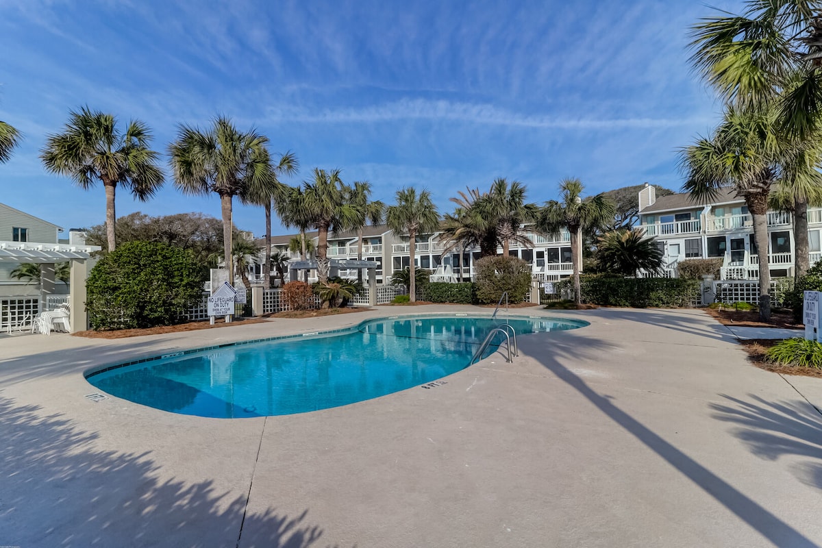 A community swimming pool is framed by lush palm trees and landscaping. The clear water reflects the blue sky above, while a surrounding concrete deck provides ample space for lounging. The pool area is bordered by townhomes in the background, enhancing the tranquil setting.