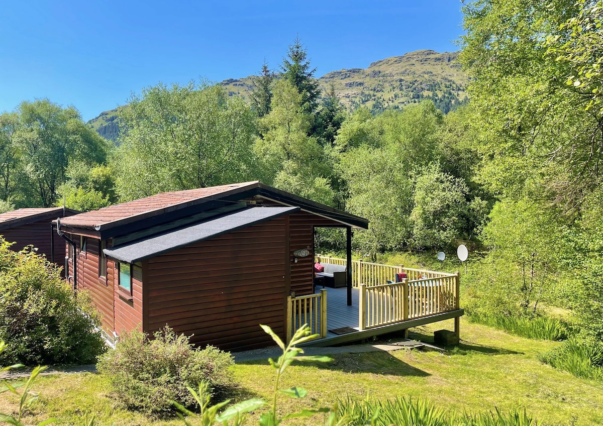 The log cabin is set among lush greenery, surrounded by trees and woodland. A covered deck extends from the cabin, offering outdoor seating. The landscape is framed by rolling hills in the background, providing a serene and natural environment.