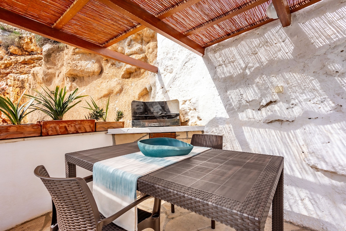An outdoor dining area features a large table with a textured surface and several chairs. A minimalistic bowl sits at the center of the table. The space is shaded by a wooden pergola, and the surrounding walls display natural stone textures. Potted plants add a touch of greenery.