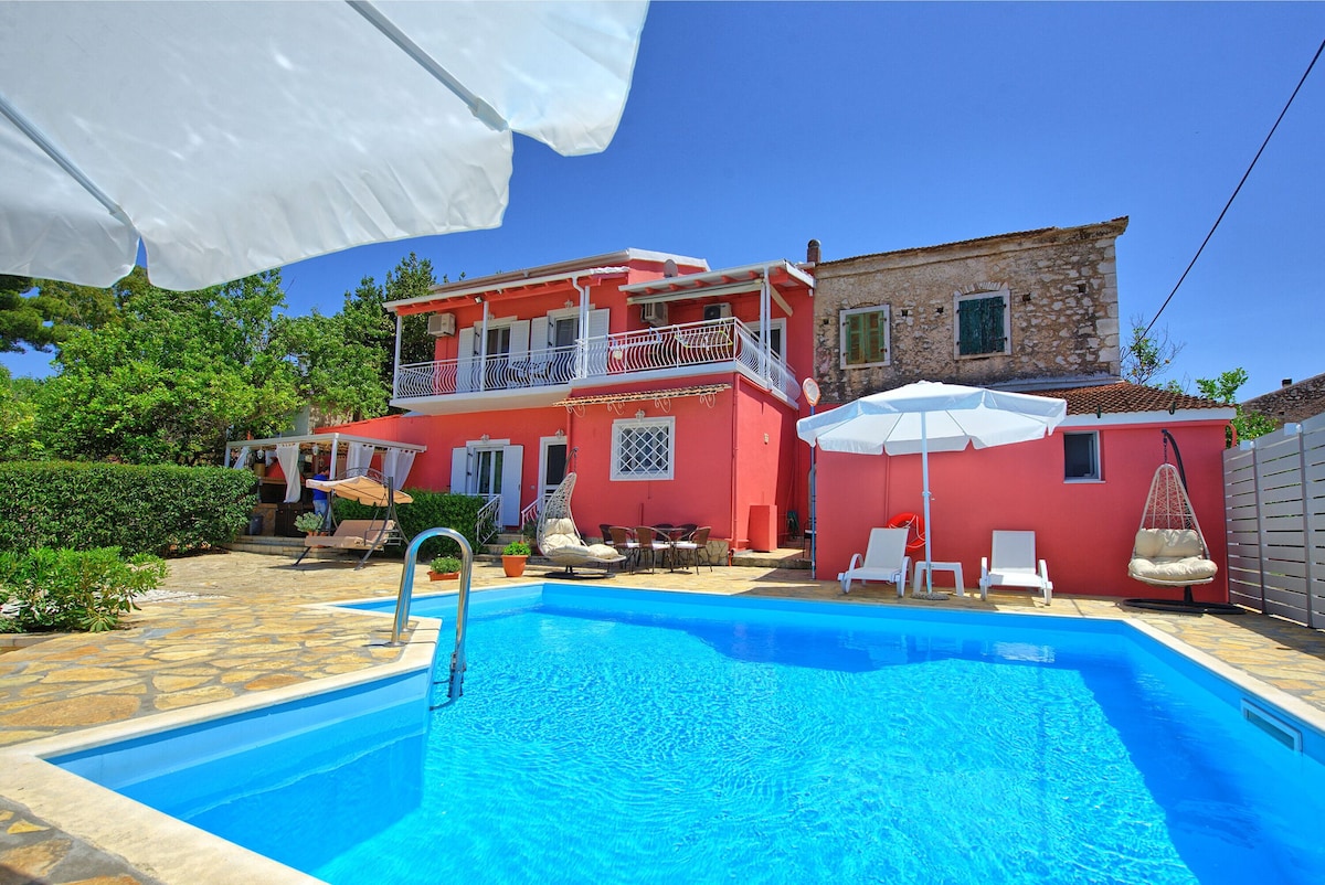 The vibrant villa is framed by lush greenery and set beside a clear blue private pool. A shaded seating area is visible next to the pool, complemented by white umbrellas. The inviting architecture showcases a mix of traditional and modern elements, enhancing the outdoor leisure space.