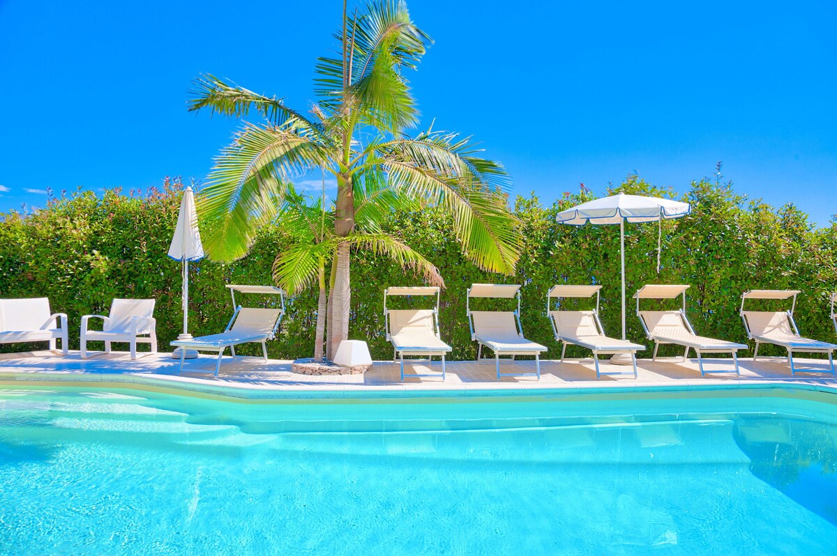 A serene swimming pool is surrounded by lounge chairs and umbrellas, creating an inviting space for relaxation. Lush greenery provides privacy, while a palm tree adds a touch of tropical charm. The bright blue sky enhances the tranquil atmosphere of the outdoor area.