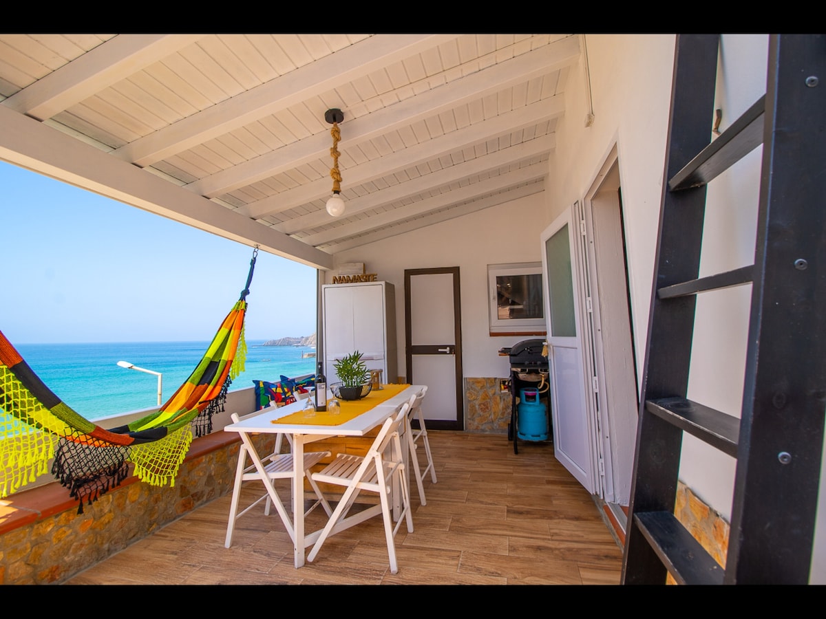 A spacious terrace features a dining table set for four and a colorful hammock inviting relaxation. The ocean view is prominent, with the tranquil blue water visible in the background. A barbecue area is present, enhancing outdoor dining options.