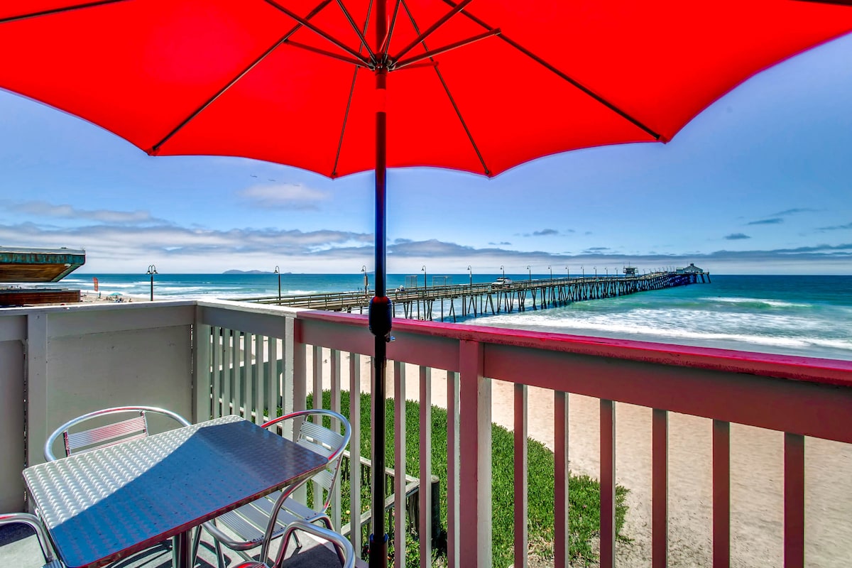 A balcony view offers an expansive ocean scene, featuring gentle waves and a fishing pier in the distance. A large red umbrella provides shade over a small table and two chairs, creating a space for relaxation and enjoyment of the coastal surroundings.