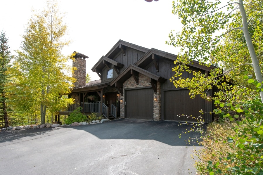 A spacious mountain home features a rustic wooden exterior with a stone chimney and two garage doors. Surrounding the home are vibrant green aspen trees, complemented by landscaped stone accents along the driveway, creating a welcoming entrance.