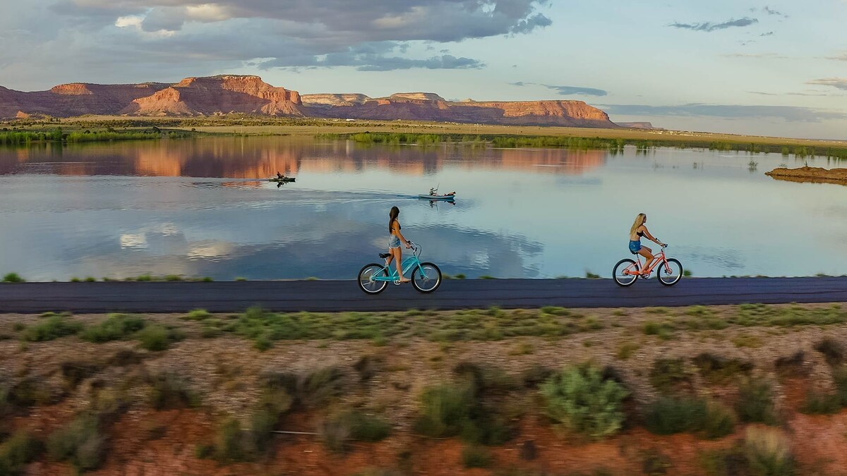 Two individuals ride bicycles along a paved path beside a calm body of water. Reflections of the surrounding red cliffs and sky are visible in the water, enhancing the serene landscape. Soft greenery borders the path, contributing to the natural ambiance.