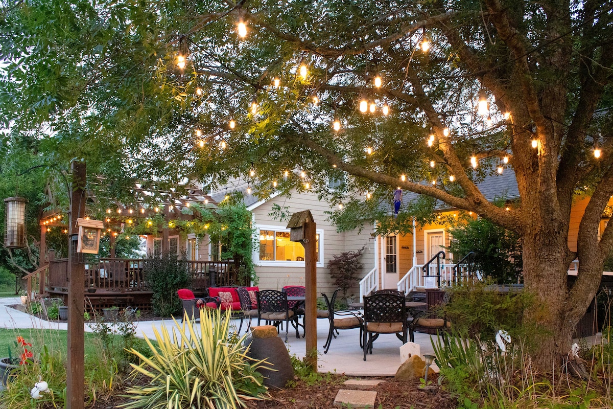 A cozy outdoor area showcases a spacious patio with gathering seating and a fire pit. Soft lighting drapes from tree branches, creating a serene ambiance. The deck and home are visible in the background, featuring a welcoming atmosphere suitable for relaxation.