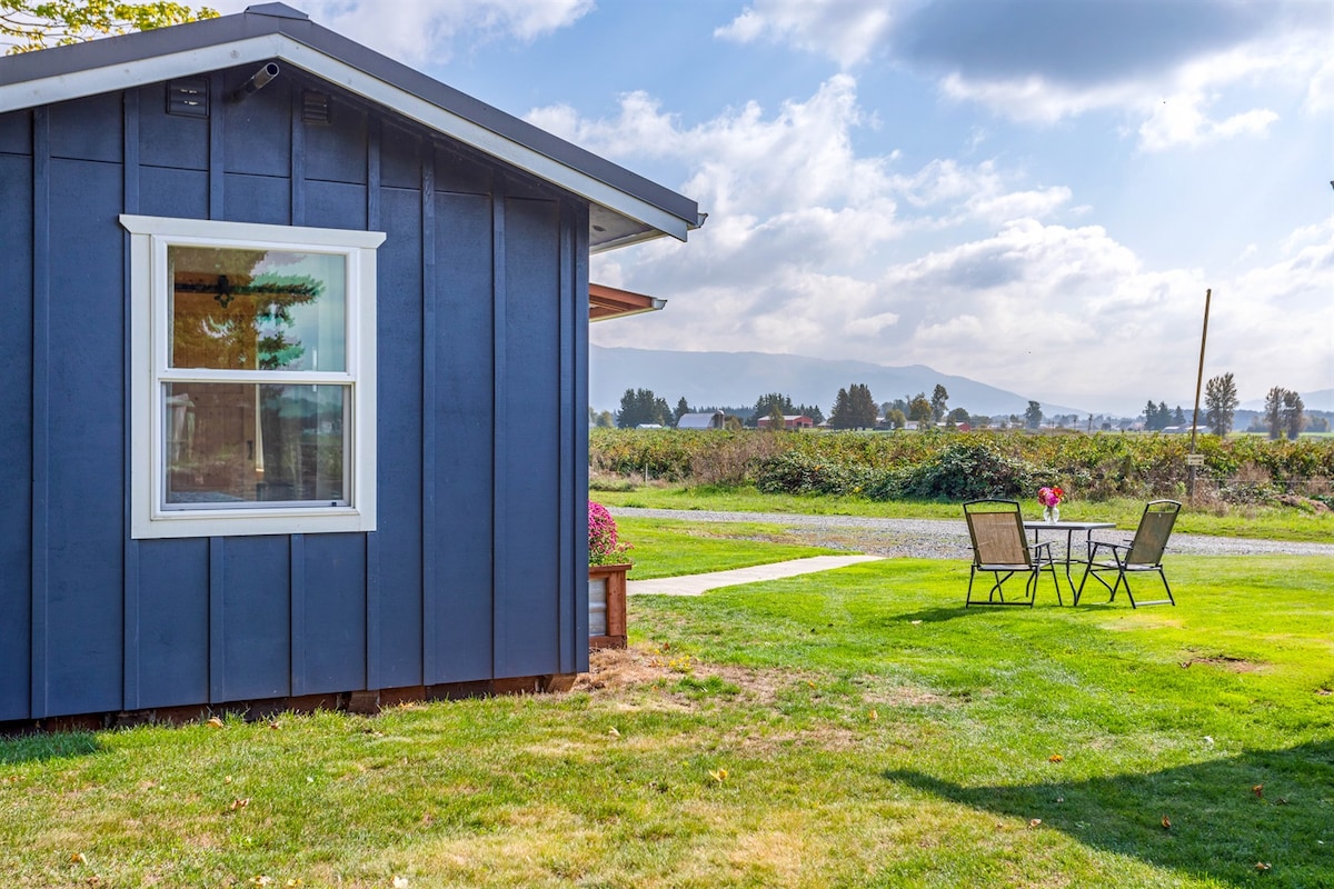 The exterior of the Little Farmhouse showcases a charming dark blue facade. A small patio area with a table and chairs is visible on the green lawn, providing a peaceful spot to enjoy expansive views of the farm and surrounding landscapes.