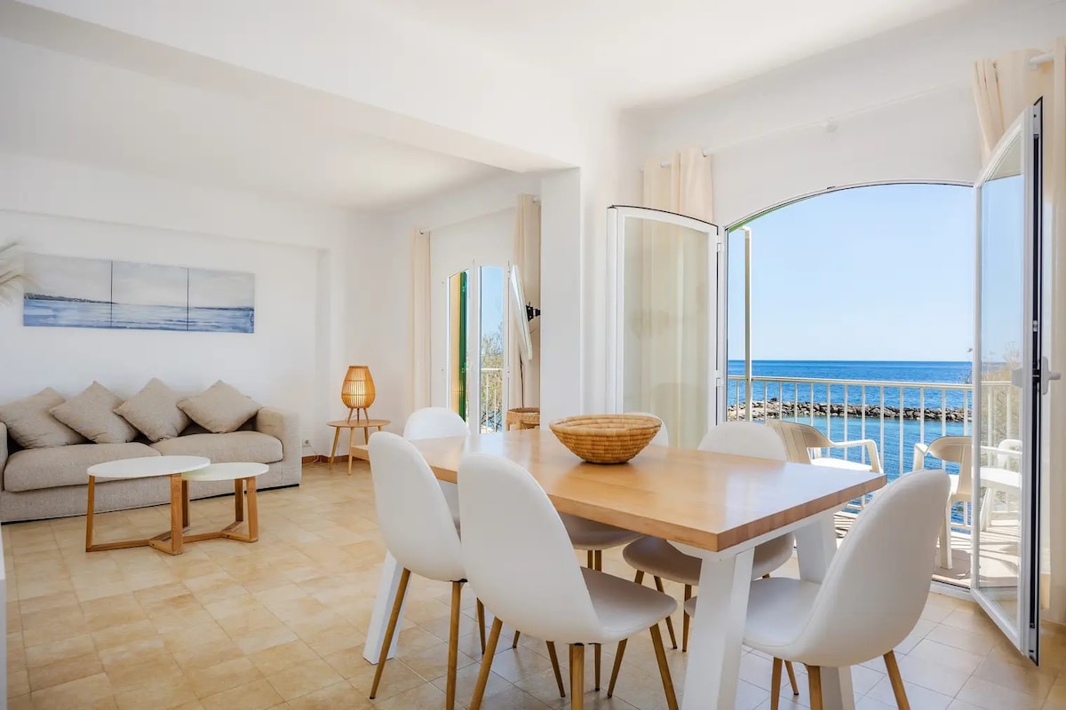 A bright dining area features a wooden table surrounded by white chairs. A woven bowl is placed at the center. An inviting sofa with neutral tones is positioned nearby, with open doors leading to a scenic view of the sea beyond.