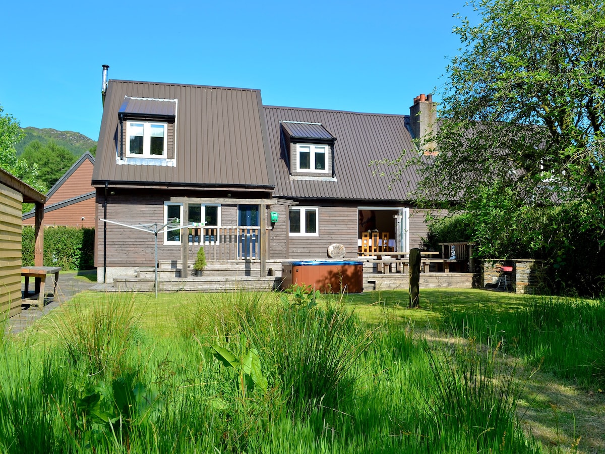 A spacious lodge is shown nestled within lush greenery, featuring a well-maintained lawn. The exterior includes a sloped roof and numerous windows. A private hot tub is visible on the patio, with outdoor furniture positioned nearby for relaxation.