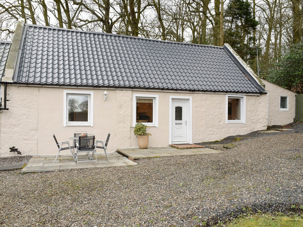 The exterior of Kinneddar Cottage features a light-colored stone facade with a dark tiled roof. A small patio area includes a table and chairs, surrounded by a gravel pathway and greenery. Windows allow natural light to fill the interior, contributing to a welcoming feel.