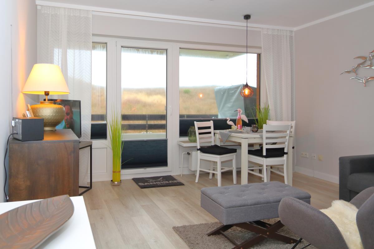 A modern living area features a light color palette and an integrated dining table for four. Large glass doors lead to a balcony, framed by sheer curtains. A stylish lamp and contemporary artwork add visual interest, while a comfortable seating arrangement invites relaxation.