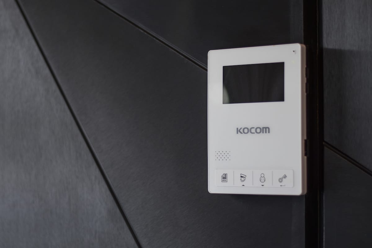 A modern intercom system is mounted on a sleek dark panel. The device features a small screen and several buttons, including icons for security and communication, emphasizing convenience and safety for guests.