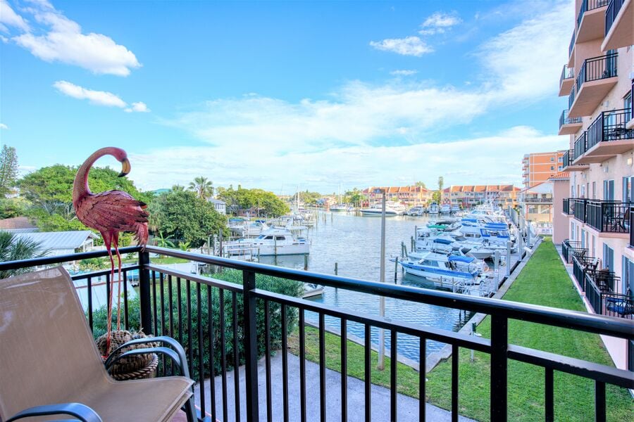 A balcony view features a metal flamingo sculpture and a scenic waterway lined with boats. Lush greenery and a manicured lawn are visible below, with buildings reflecting the blue sky in the background. The overall setting conveys a peaceful outdoor space.