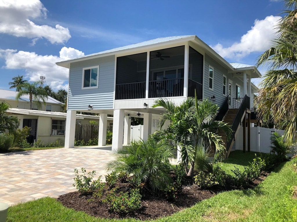 A modern two-story home is surrounded by lush greenery and well-maintained landscaping. The exterior features light blue siding and a large covered porch with a ceiling fan. A paved driveway leads up to the entrance, framed by palm trees and flowering bushes.