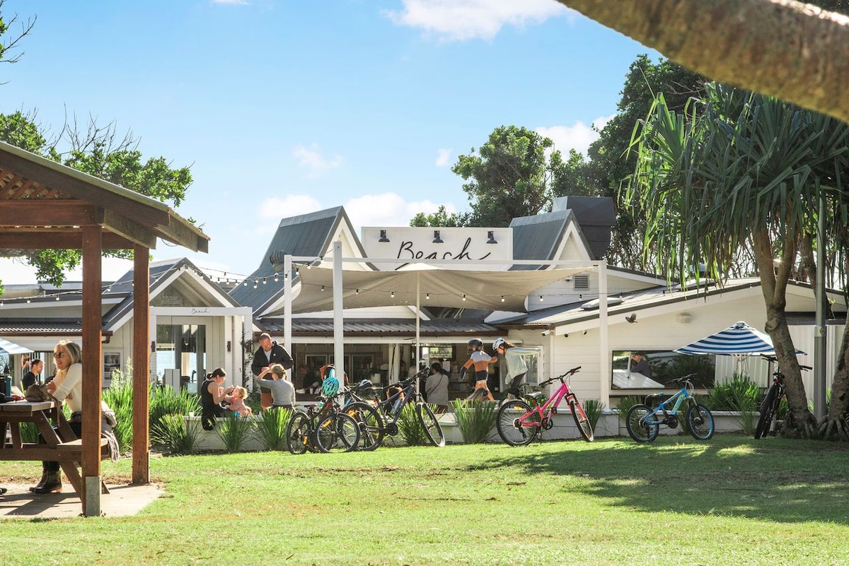A vibrant café setting features outdoor seating under shaded awnings. Bicycles are parked nearby, and patrons enjoy their meals in a relaxed atmosphere. Lush green grass and palm trees frame the scene, with a clear blue sky overhead enhancing the inviting environment.