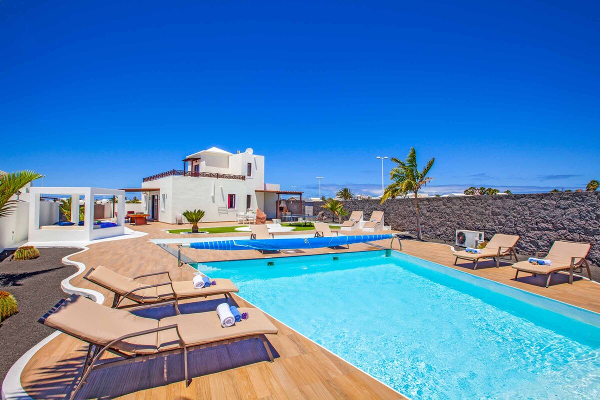 The outdoor area features a large freeform swimming pool surrounded by a wooden deck and lined with sunbeds. Lush greenery adds a natural touch, and the villa is visible in the background, contributing to the spacious and inviting environment.