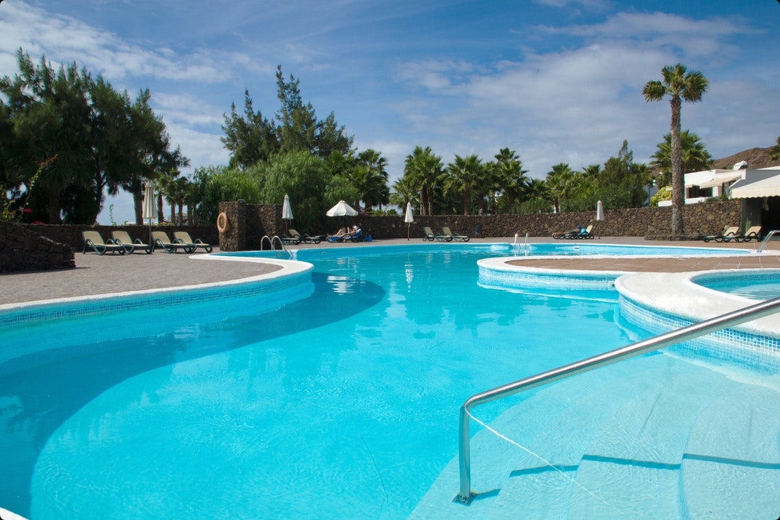 A shared outdoor swimming pool is shown, featuring shaded areas with loungers surrounding the water. Lush palm trees and well-maintained gardens contribute to the tranquil environment. The pool design includes separate areas for adults and children.