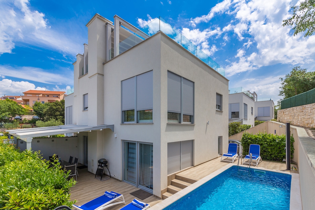 The modern two-story building is surrounded by greenery, featuring a private pool alongside a spacious deck area. Sun loungers are positioned near the pool, while a covered outdoor space provides shaded seating. Large windows reflect the bright blue sky, creating an airy and inviting exterior.