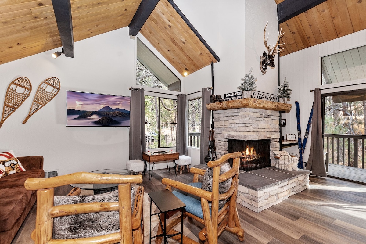 A welcoming living area features a large stone fireplace with a wood mantel, providing a focal point in the space. Vaulted ceilings add to the room's openness. Two leather accent chairs are positioned beside a small side table, and a flat-screen TV is mounted on the wall.