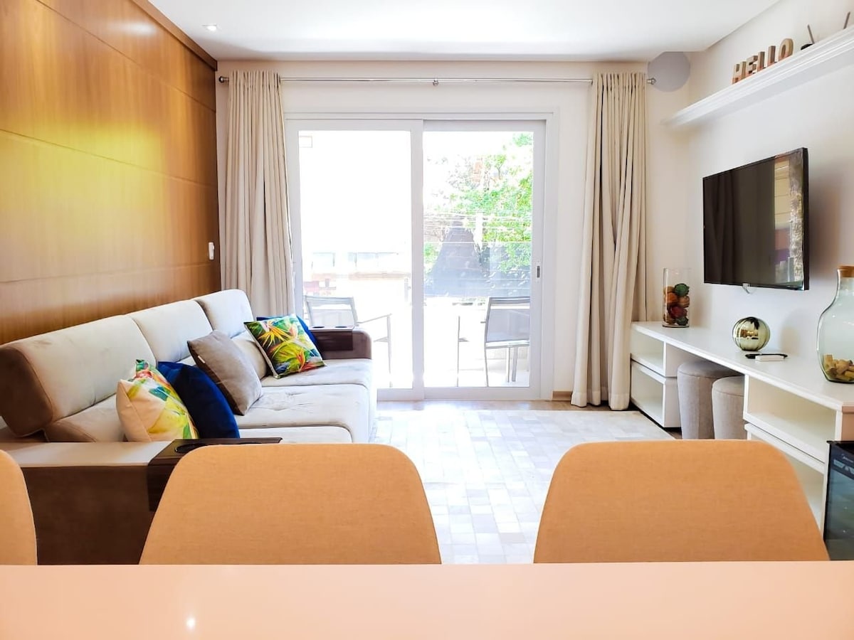 A living area features a spacious sofa adorned with colorful cushions, positioned in front of a wall-mounted television. Natural light fills the room through sliding glass doors that lead to an outdoor space. Light-colored walls and soft curtains create a bright and airy atmosphere.